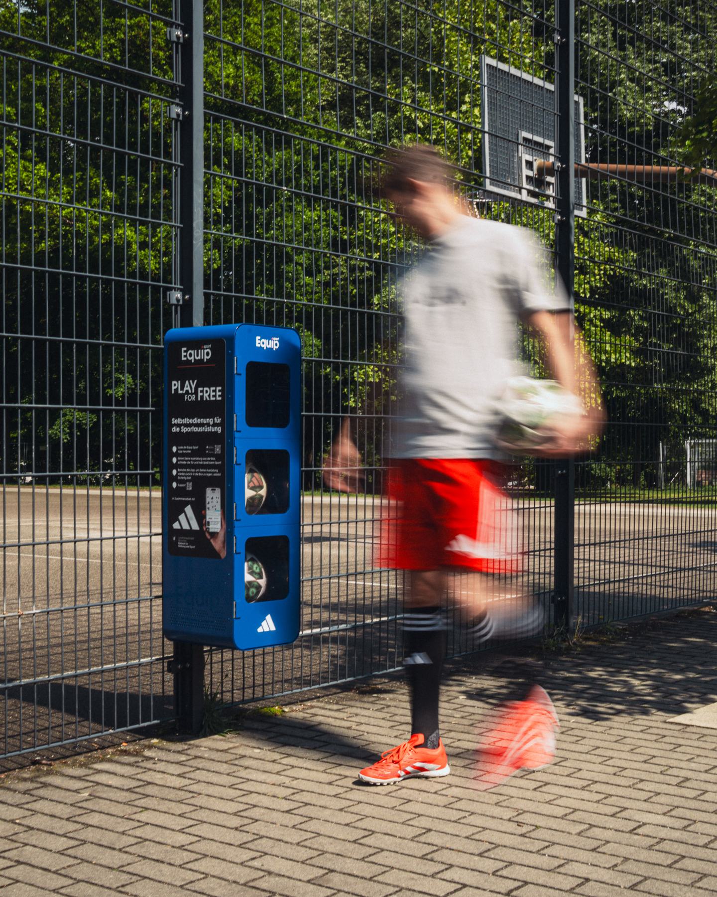 A player walks past an Equip Sport station at a Munich street court, the ball under his arm.